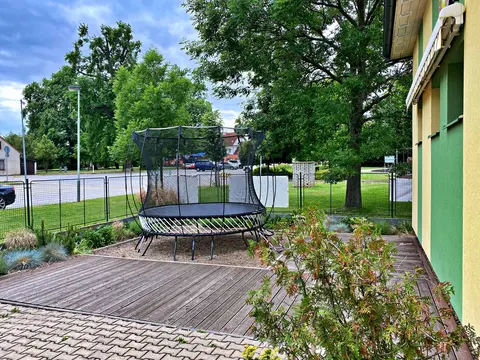 Venkovní trampolína s ochrannou sítí na dřevěné terase vedle budovy a stromů
