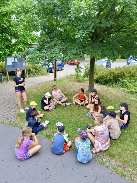 Skupina dětí sedí v kruhu na trávě pod stromem s vedoucí
