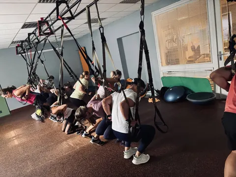 Plný sál cvičenců ve dřepu v TRX popruzích
